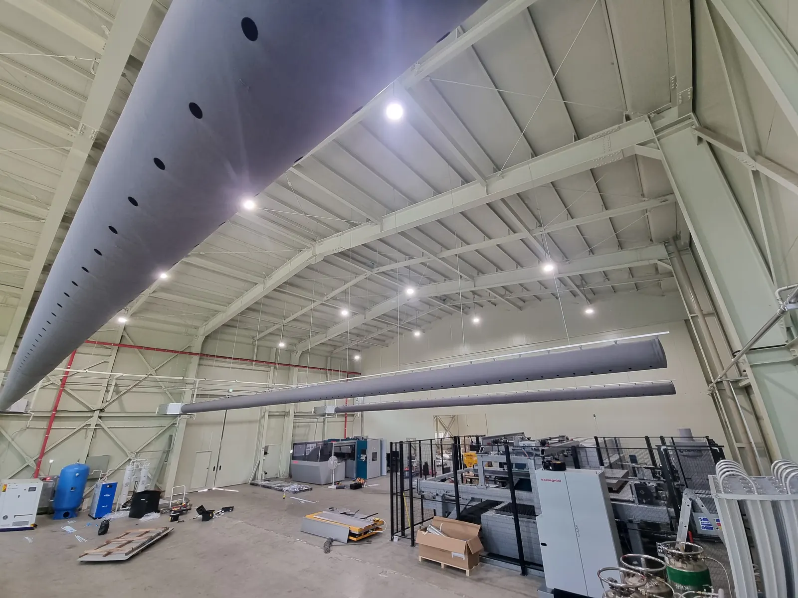 Large grey round fabric duct with a row of nozzles hung under the steel roof of a warehouse, with two more ducts in the background