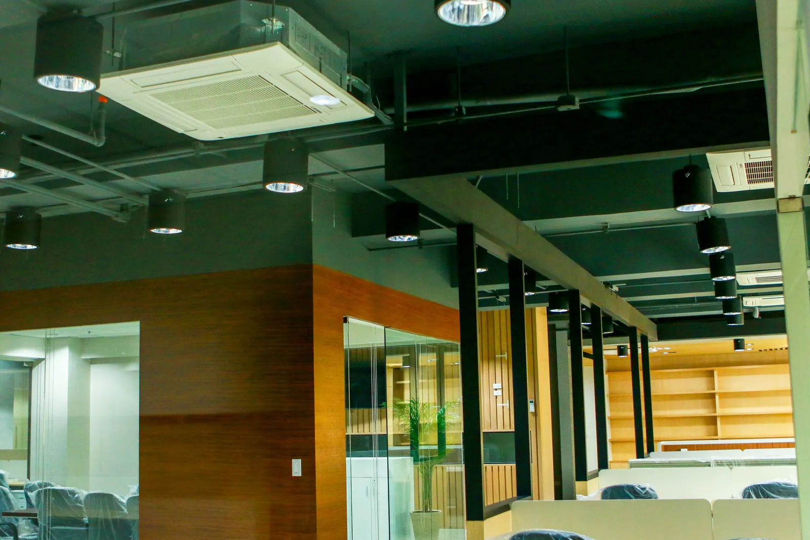 Ceiling cassette aircon units mounted in a dark exposed ceiling of an office, between pendant lights, with a glass-walled room below