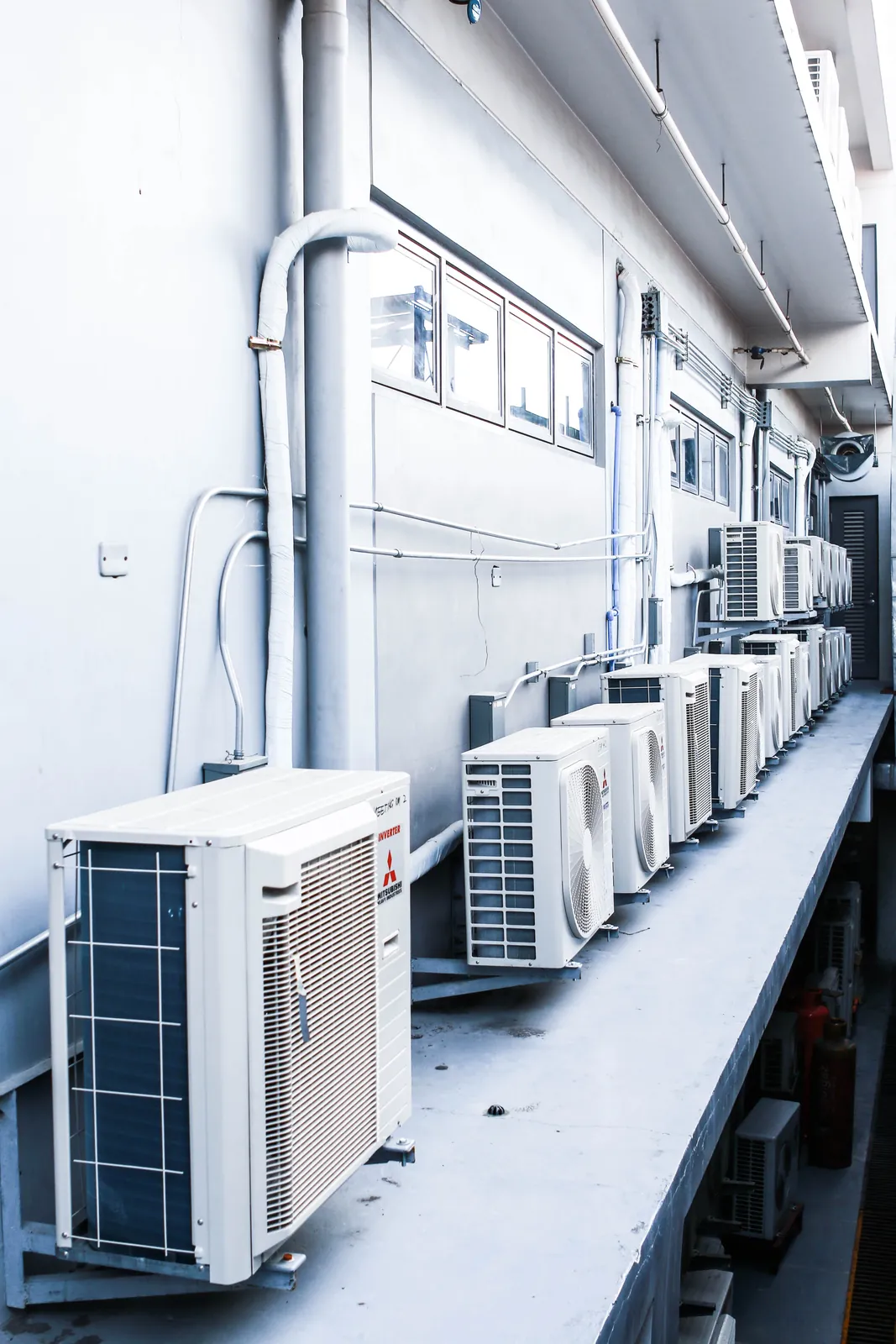 Row of inverter aircon outdoor units on a ledge along a building wall, with insulated refrigerant piping and drain lines