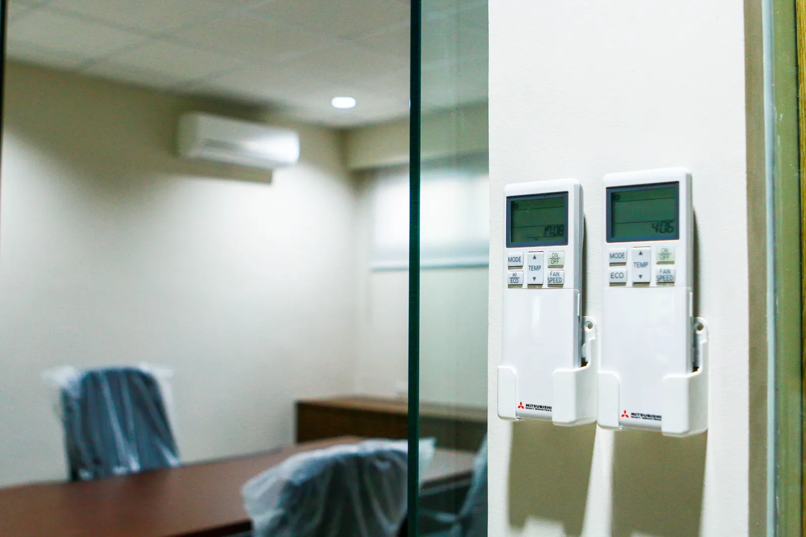 Two aircon remote controllers in a wall holder next to a glass meeting room with a wall-mounted split-type indoor unit